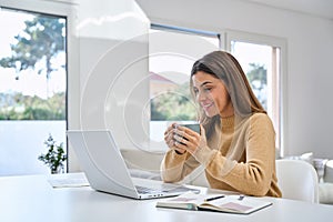 Smiling happy middle aged woman drinking coffee looking at computer at home.