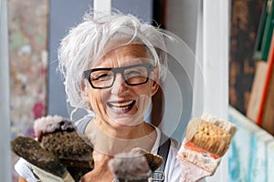 Smiling happy older woman portrait, proud artist, in her fifties with grey hair and black glasses and many paintbrushes