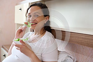 Smiling happy positive pregnant woman eats a healthy salad with fresh raw vegetables and herbs, looking at camera