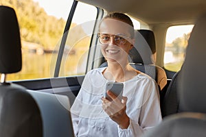 Smiling happy positive woman is using a smart phone and smiling while sitting on back seat in car using mobile phone looking at