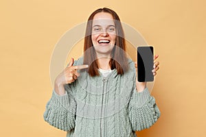 Smiling happy positivepregnant woman wearing knitted warm sweater standing isolated over beige background pointing at smart phone
