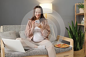 Smiling happy pregnant woman sitting on sofa having break of her online work using smartphone for browsing web pages about