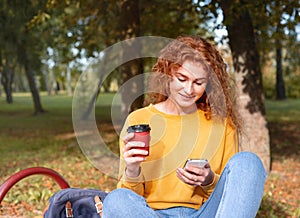 Smiling happy red hair student girl having a break with coffee outside in autumn park