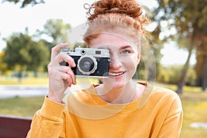 Smiling happy red hair student girl taking picture with old vintage camera outside in autumn park