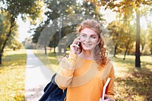 Smiling happy red hair student girl talking on the phone outside in autumn park