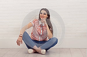 Smiling happy. Relaxed happy young woman sat cross-legged on floor, touching chin, looking at camera, isolated white brick wall