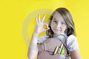 Smiling happy schoolgirl. cute caucasian girl with blond hair holds backpack with school supplies and shows OK sign on