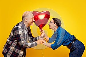 Smiling, happy, senior couple, man and woman holding balloon in heart shape, dating, celebrating against yellow