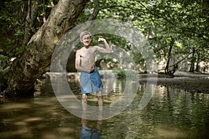 Smiling happy teenage boy having fun by posing like bodybuilder in shallow water in sunny forest in mediterranean country in