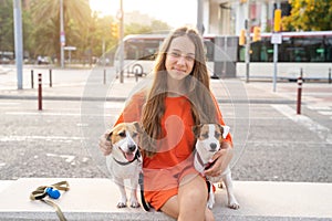 Smiling happy teenage girl in orange clothes hugging cute pets Jack Russells sitting on the bench