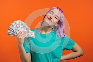 Smiling happy woman with cash money - USD currency dollars banknotes on orange wall. Girl with violet dyed hairstyle