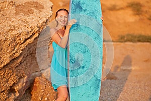 Smiling happy woman in dress with surfboard on mountain background. Surfing sport, summer time and holiday trip concept