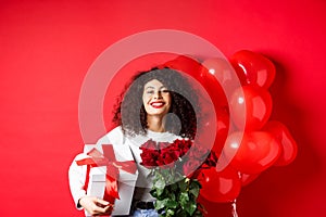 Smiling happy woman holding box with gift and red roses from boyfriend, celebrating Valentines day, standing near