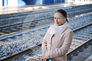 Smiling happy woman in warm beige coat standing outdoor on the platform of a railway station, waiting to board the train