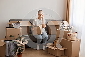 Smiling happy woman wearing white t-shirt and jeans sitting on cough in a new apartment after moving, posing with cardboard