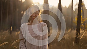 smiling happy woman in white light dress walking in autumn forest between golden spikelets of tall dry grass and pine