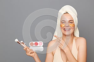 Smiling happy woman wrapped in towel standing cosmetics brushes and palette of eye shadows in hands, pointing away at