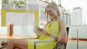Smiling happy woman in yellow dress sitting in chair near swimming pool. Using her smartphone