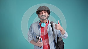 Smiling happy young adult arab or indian man student standing isolated on blue background, showing thumbs up like hand