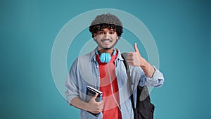 Smiling happy young adult arab or indian man student standing isolated on blue background, showing thumbs up like hand