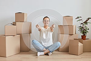 Smiling happy young adult woman wearing white T-shirt sitting on the floor near cardboard boxes with stuff, posing in lotus pose