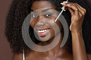 Smiling happy young African black woman applying serum oil on facial skin.