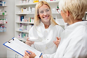 Smiling happy young blond-haired pharmacist holding a box of medications, consulting with her older colleague