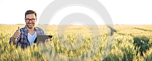 Smiling happy young farmer or agronomist using a tablet in a wheat field. Showing thumbs-up and looking directly at camera