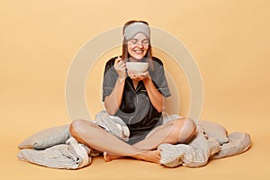 Smiling happy young girl sitting floor with blanket wearing black pajama and sleep eye mask holding plate in hands isolated over