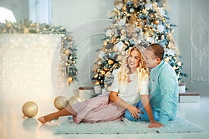 Smiling happy young married couple wife and husband sitting near christmas tree with christmas lights around. Boyfriend and