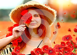 Smiling and happy young red-haired  woman in a black dress and a hat holding red poppies near the face. Beautiful poppy field at