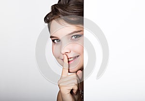 Smiling happy young woman standing behind and leaning on a white blank billboard or placard, expresses different