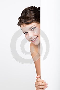 Smiling happy young woman standing behind and leaning on a white blank billboard or placard, expresses different