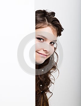 Smiling happy young woman standing behind and leaning on a white blank billboard or placard, expresses different