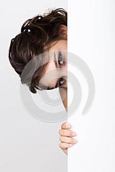 Smiling happy young woman standing behind and leaning on a white blank billboard or placard, expresses different