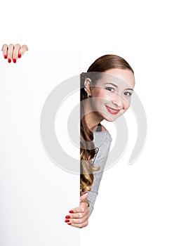 Smiling happy young woman standing behind and leaning on a white blank billboard or placard, expresses different