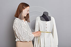 Smiling hard working female sewer standing near mannequin with new dress isolated over gray background measuring new dress