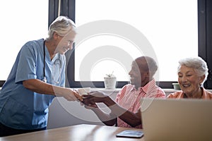 Smiling healthcare worker serving coffee to senior man sitting by friend