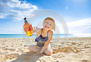 Smiling healthy child in swimwear on seacoast showing lotion