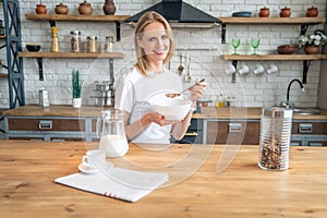 Smiling healthy woman eating corn flakes cereal while sitting and having breakfast at the kitchen table