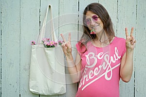 Smiling hippie woman in pink t-shirt with Be Cheerful inscription and pink round glasses, made Peace sign standing near blank