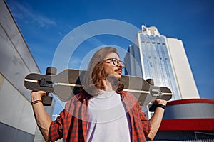 Smiling hipster man with longboard on shoulder over city background