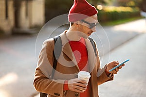 Smiling hipster man wear sunglasses using cellphone standing at the street drinking takeaway coffee
