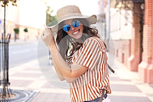 Smiling hipster trendy girl posing at the city summer street,holding hat