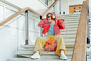 Smiling hipster young woman in bright clothes, sunglasses with backpack bag and wireless headphones listening to music