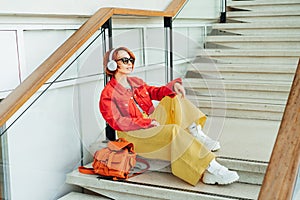 Smiling hipster young woman in bright clothes, sunglasses with backpack bag and wireless headphones listening to music