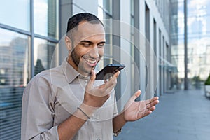 Smiling hispanic businessman recording voice message, happy man outside office building reading commands on smartphone