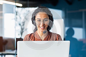 Smiling hispanic businesswoman working inside office with laptop and headset for video call, woman sitting at workplace