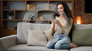 Smiling Hispanic Caucasian 30s woman housewife girl sitting on couch cozy sofa at home using smartphone for ordering