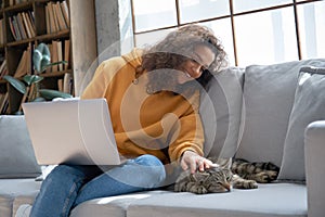 Smiling hispanic girl using laptop playing with cat sitting on couch at home.